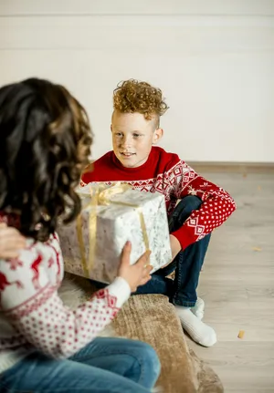 A mum hands a large golden gift to her son, sitting on the floor in a Christmas jumper