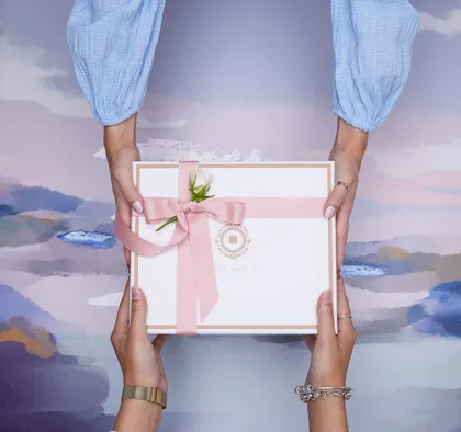 Two hands exchanging a ribboned gift over a painted lilac background