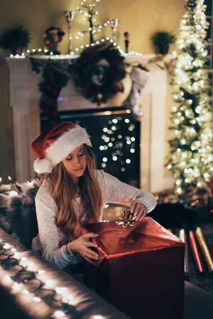 A woman in a Santa hat wraps a big red gift by the tree